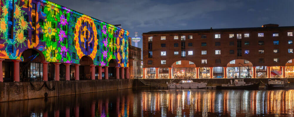 Colourful patterns are projected on to the side of a building in Liverpool's docks area in the night during the installation, Ukrainian Dreams. Lights reflect off the water in the foreground.