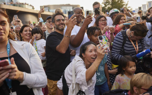 A large crowd stands close together outdoors in the sunshine during a GDIF performance. People of different ages smile and look upward. Many hold up phones to film and take photos. The scene feels lively and full of excitement.
