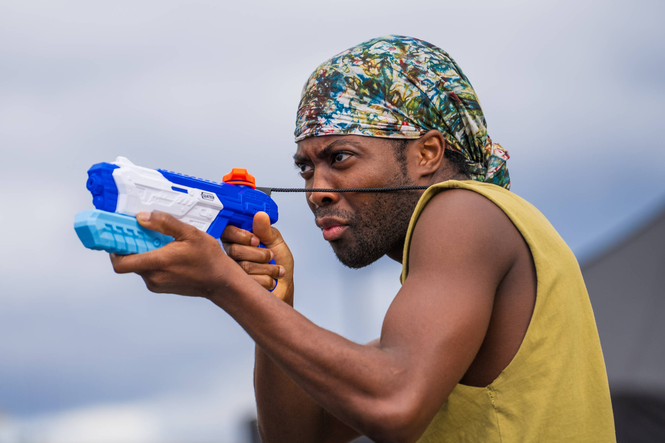 A dancer wearing a patterned headscarf and yellow sleeveless shirt aims a brightly colored blue and white toy water gun, pulling back the elastic cord in a focused pose against a cloudy sky background.