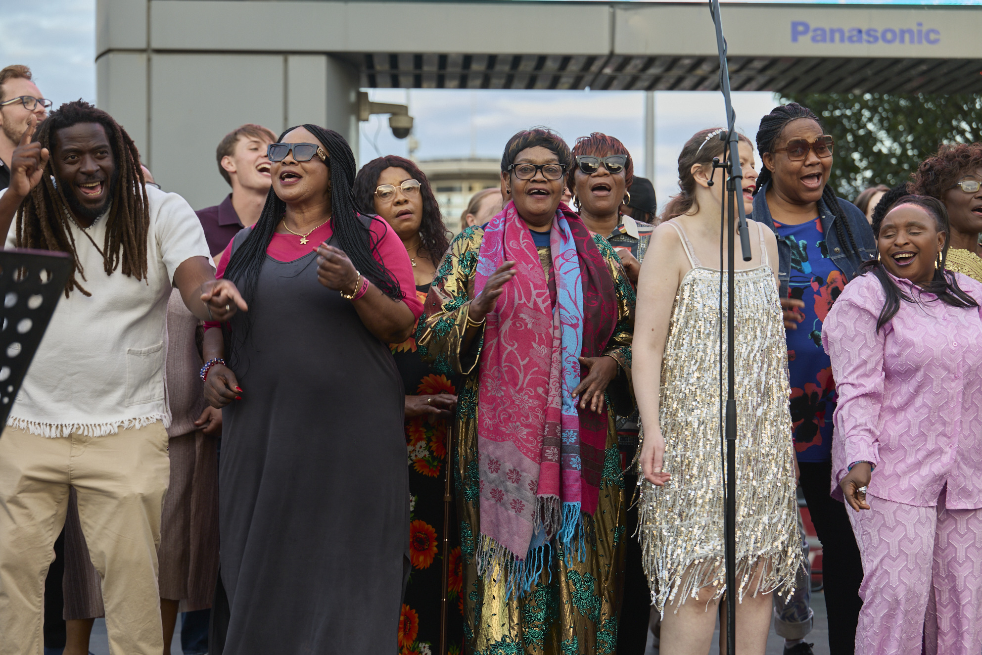 Members of the Citizens of the World Choir sing energetically outdoors in Woolwich. The diverse group, dressed in bright and patterned outfits, perform joyfully together, some smiling and clapping beneath a clear sky.
