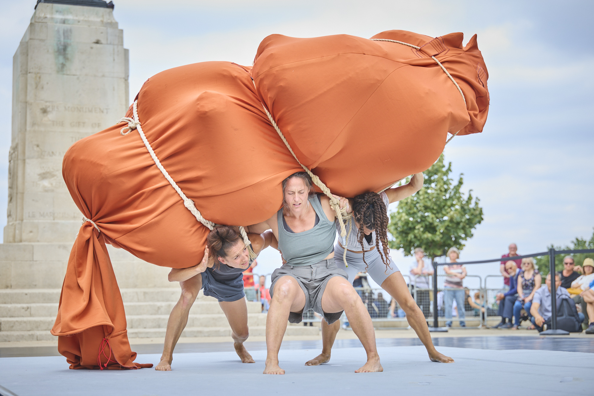 Three female performers strain together to lift and balance a huge orange fabric sculpture during a contemporary outdoor theatre piece at Greenwich Fair, while onlookers watch nearby.