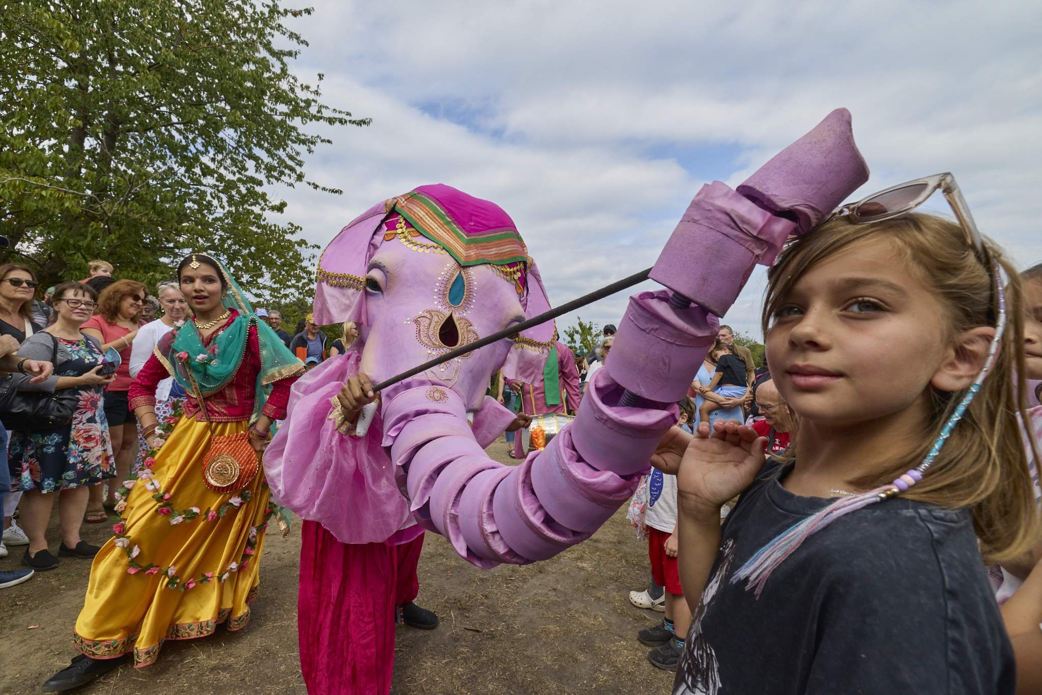 A performer in a bright traditional costume walks beside a person in a large pink elephant puppet costume, interacting playfully with children in the audience at Greenwich Fair.
