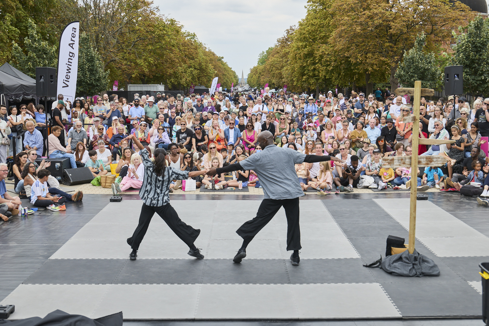 Two dancers perform a duet on an outdoor stage at Greenwich Fair, reaching towards each other as a large crowd of families and spectators watch on a tree-lined avenue.