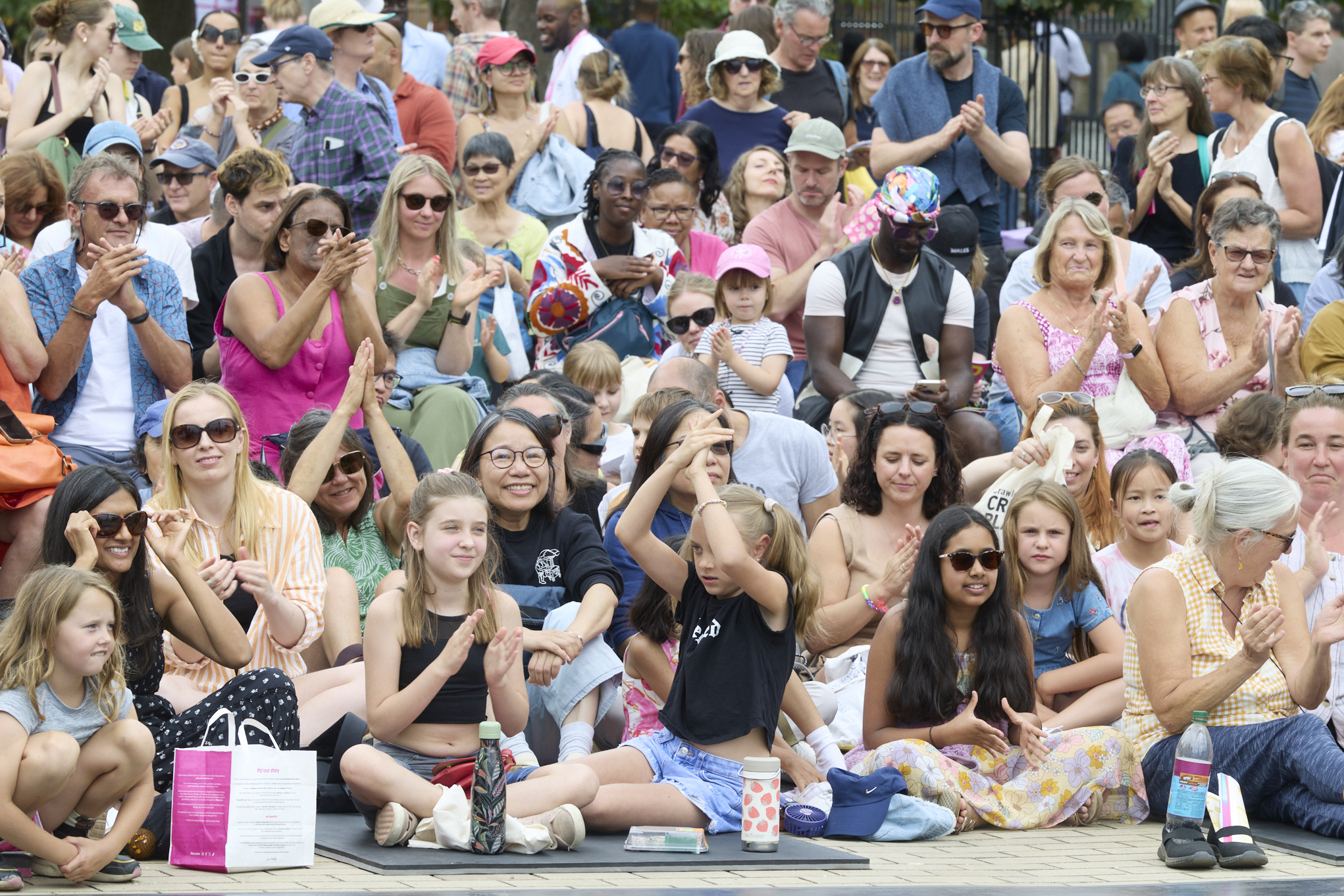 A large, diverse crowd of families and friends sit outdoors at Greenwich Fair, smiling, clapping, and enjoying a live performance. Children sit at the front, joining in the applause.