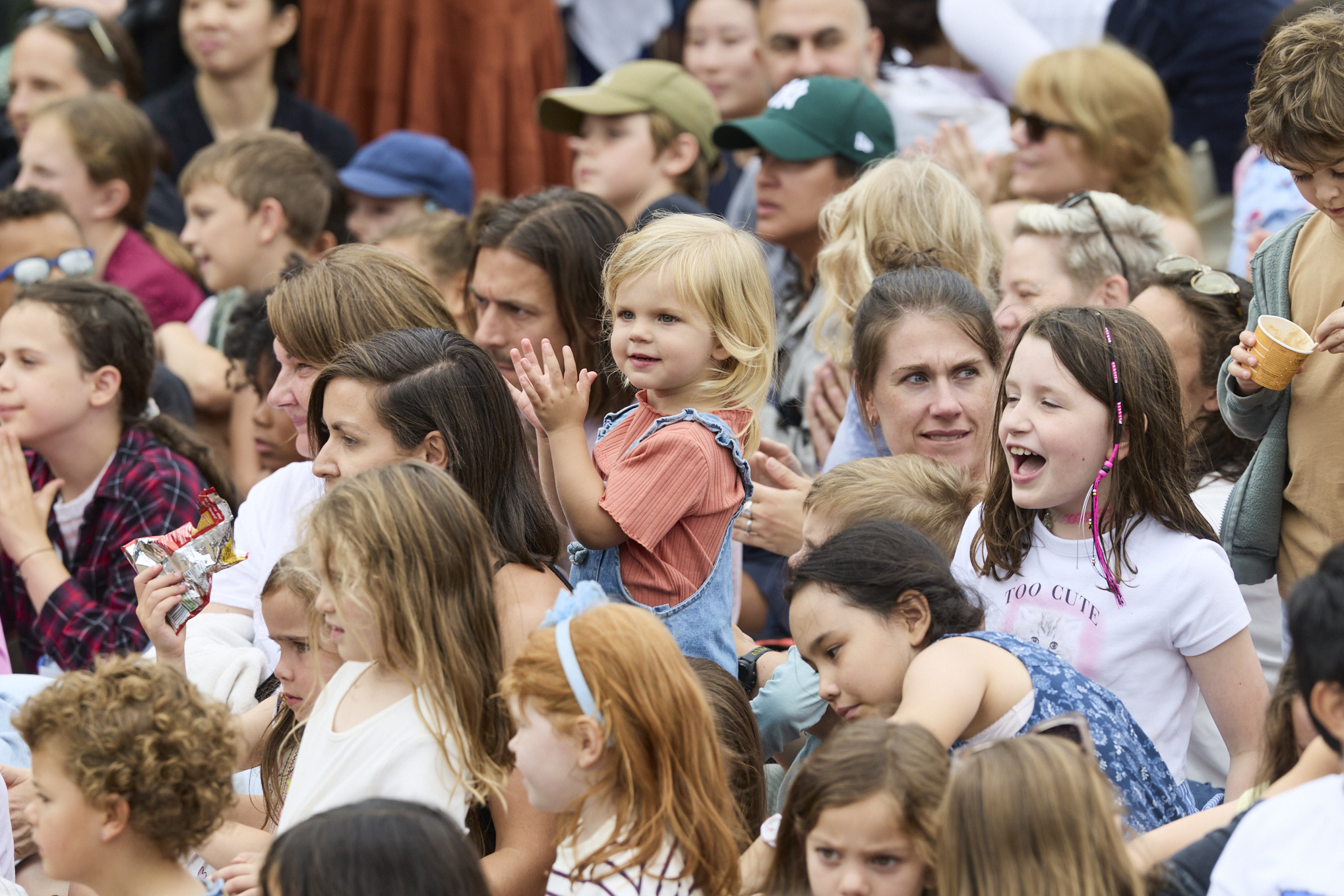 A group of children and parents watch a performance at Greenwich Fair. A young child in a red top and denim dungarees stands and claps, surrounded by smiling faces and people enjoying the show.