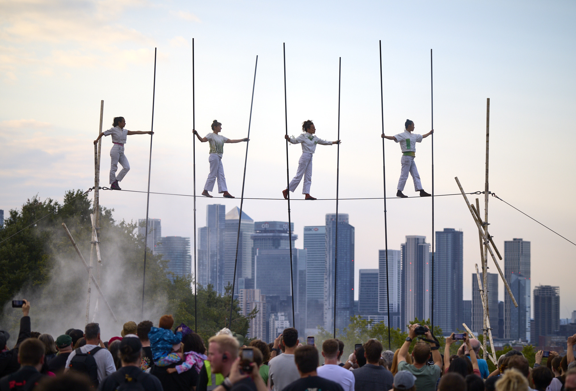 Four female tightrope walkers balance on a high wire with long poles, performing above a crowd gathered on Greenwich Park Hill. The London skyline stretches behind them under a soft evening sky.