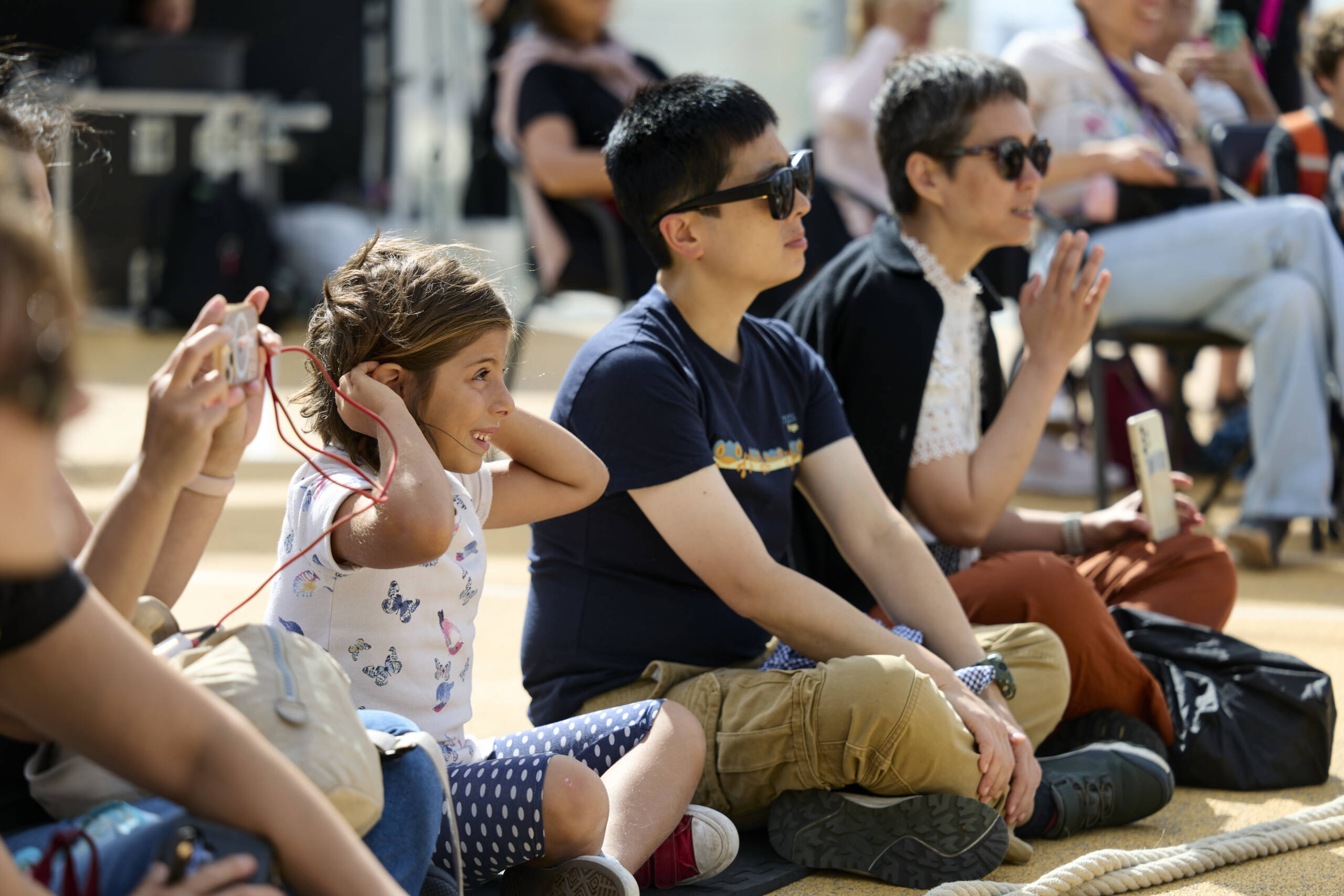 Audience members sit in the sunshine watching an outdoor performance. A young girl covers her ears beside adults who are smiling, clapping, and taking photos.