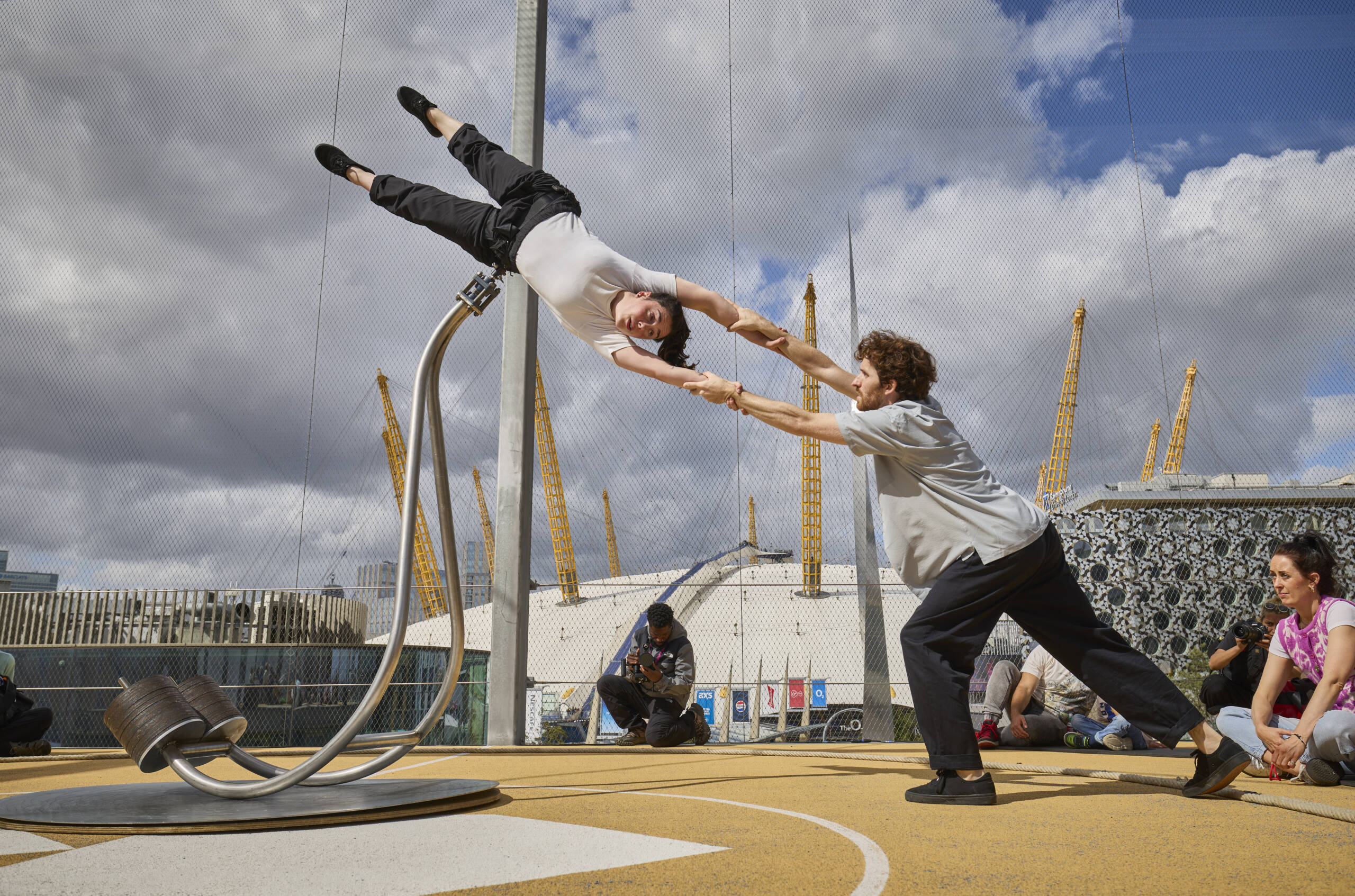 Two performers interact on a curved metal structure during The Scale, one suspended horizontally while the other supports them by the hands. The O2 Arena’s yellow masts rise in the background beneath dramatic clouds.