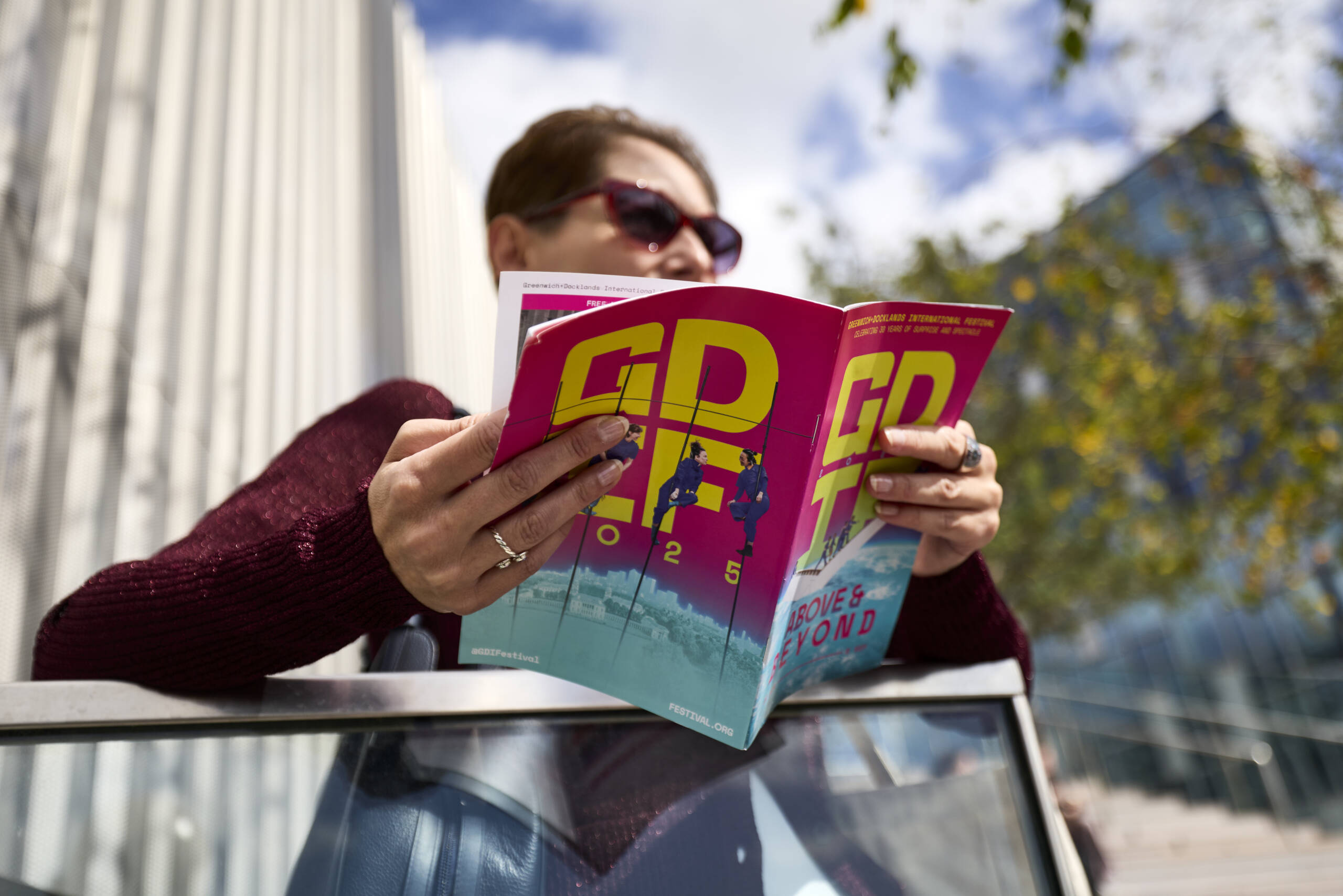 A person wearing sunglasses and a red jumper reads a colourful GDIF 2025 festival programme outdoors, with modern buildings and trees visible behind them.