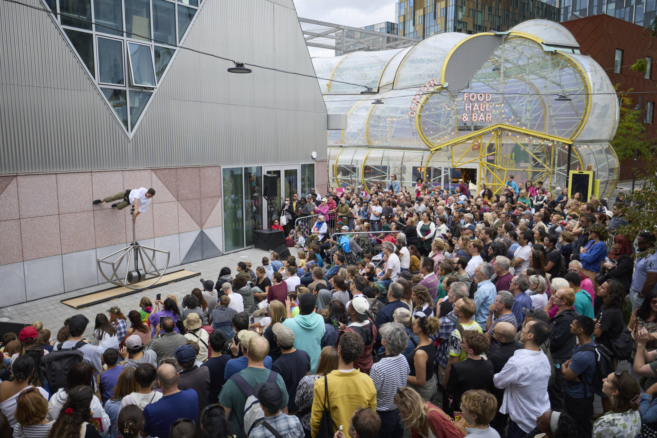 A large crowd gathers outside a modern building to watch a circus performer suspended horizontally from a metal structure in Walking on the Wall, with the entrance to a food hall and bar visible in the background.