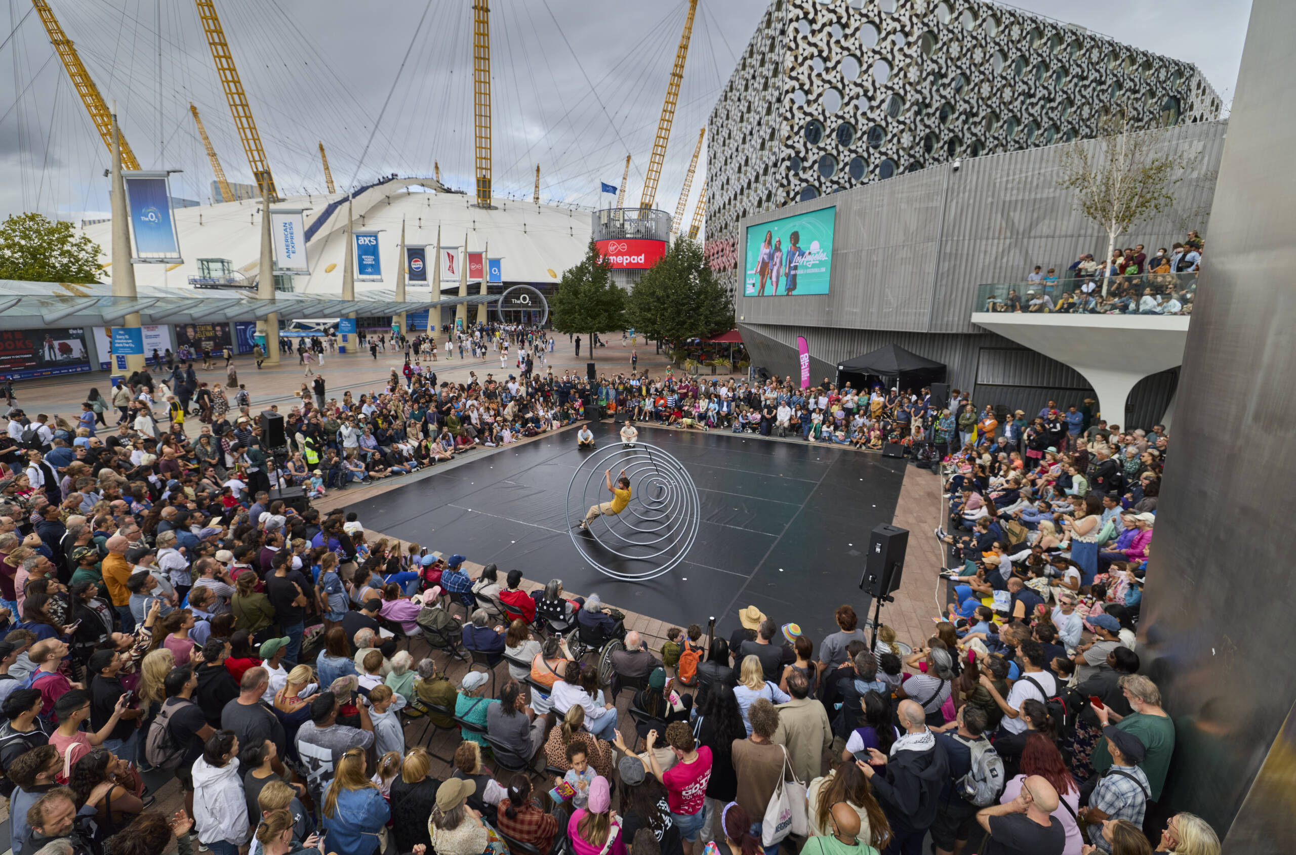 A large audience gathers in front of Greenwich Peninsula to watch Ripple, a circus performance featuring a giant metal spiral. Three performers move across the circular structure on an outdoor stage surrounded by crowds.