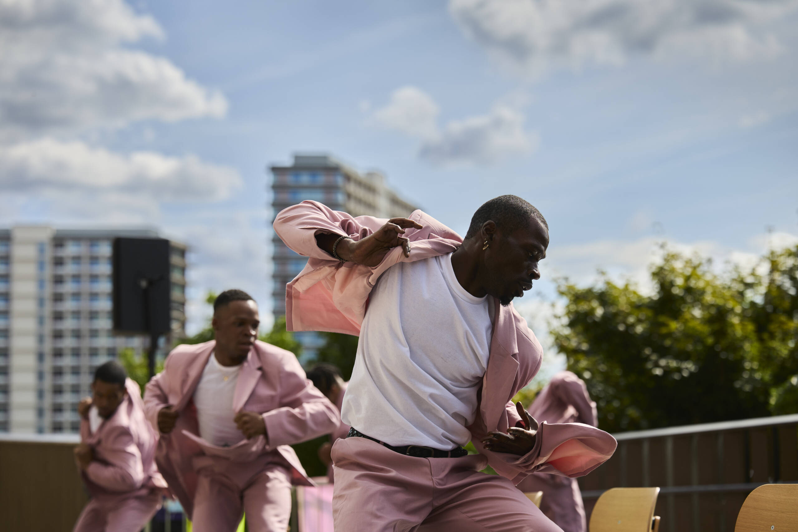 A group of male dancers in pink suits and white T-shirts perform a powerful, synchronized routine outdoors. The main dancer in the foreground moves energetically, captured mid-motion against a backdrop of trees and tall city buildings.