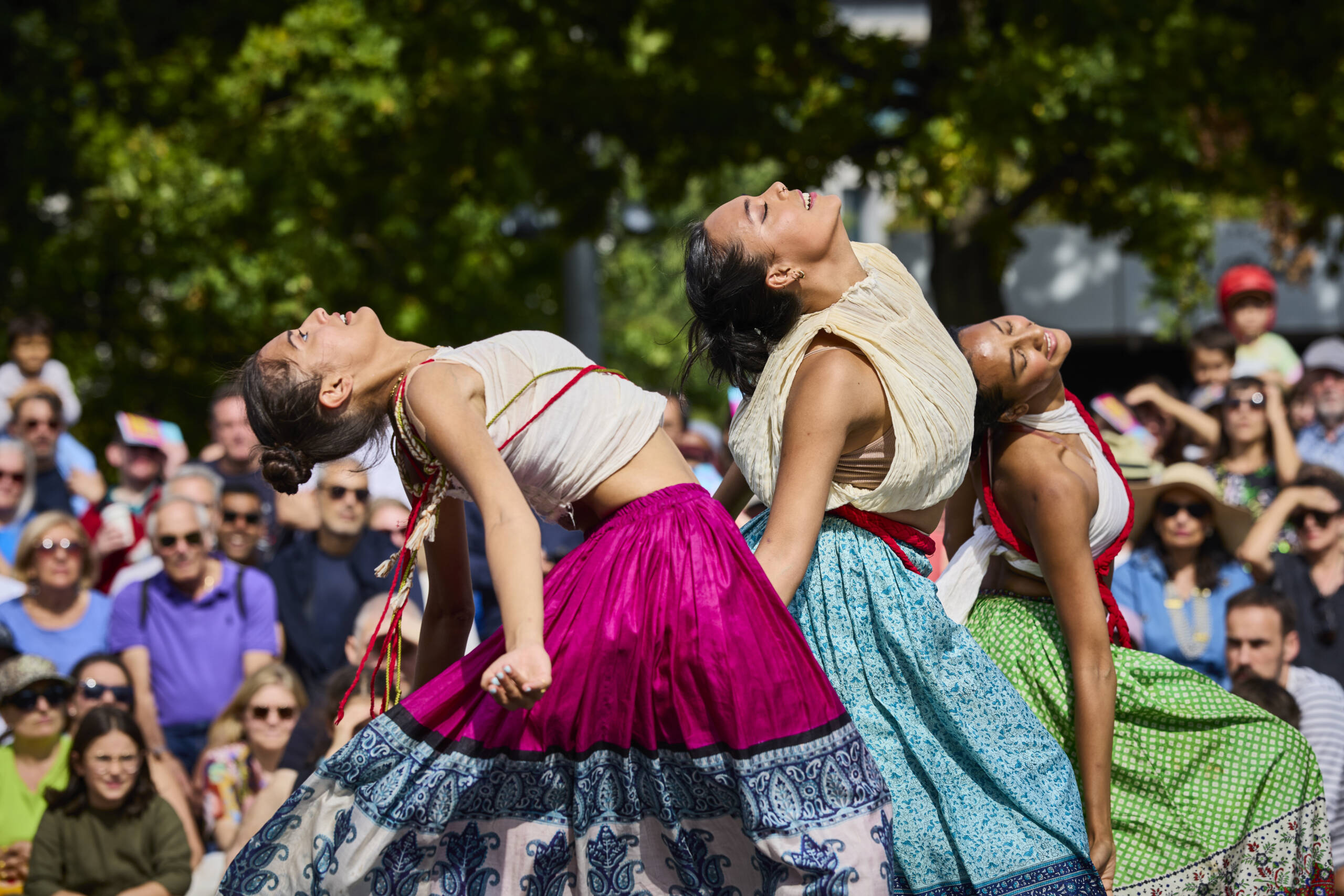 Three dancers wearing bright flowing skirts in pink, blue, and green lean back in unison, faces lifted joyfully toward the sun, as an outdoor crowd watches behind them.
