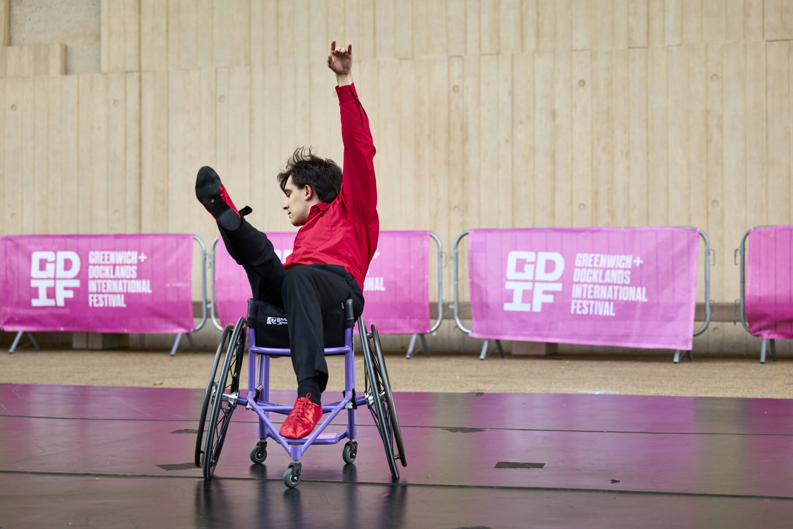 A male dancer in a red shirt performs in a wheelchair, raising one arm and one leg high in the air against a backdrop of pink Greenwich + Docklands International Festival banners.