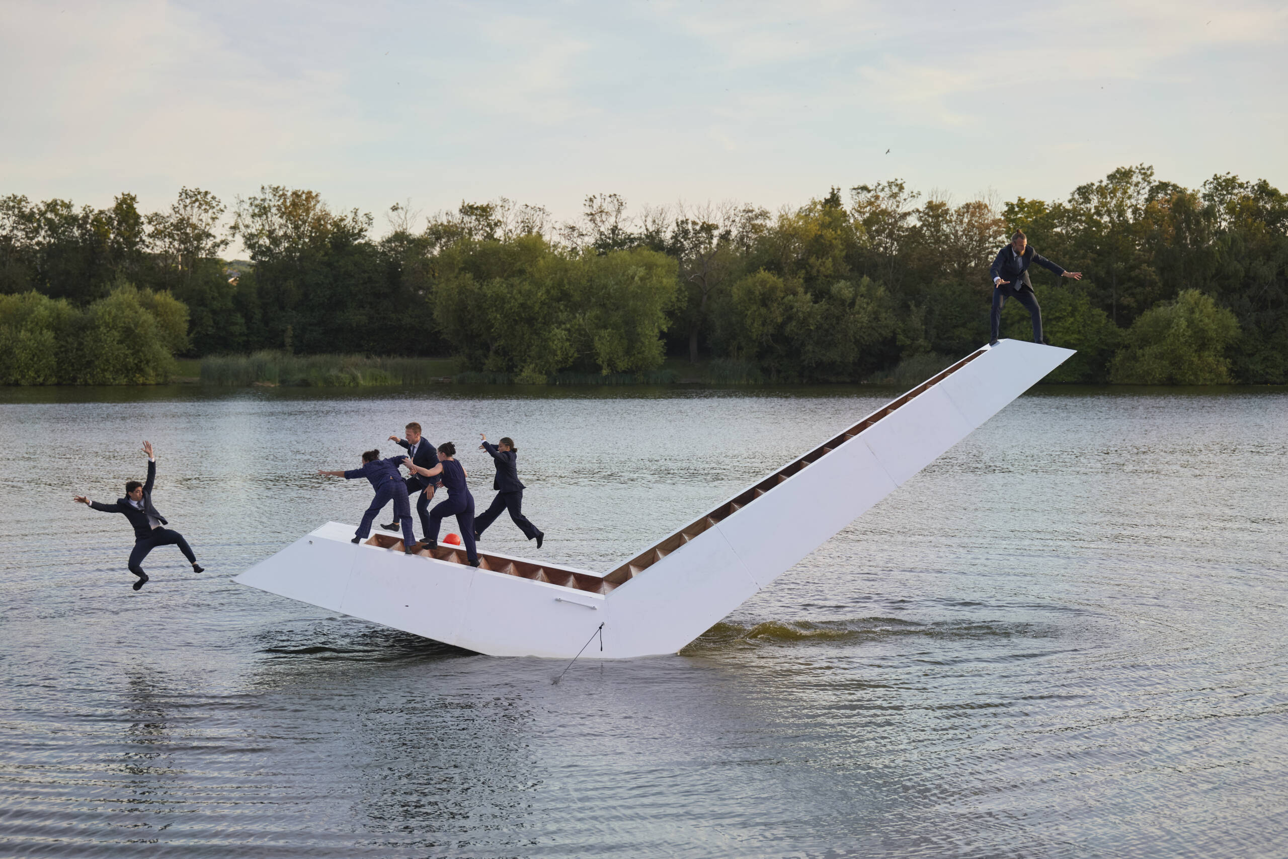 Performers in dark suits balance and leap on a large white V-shaped floating structure during a lakeside performance, with one person mid-fall into the water as others maintain their balance.