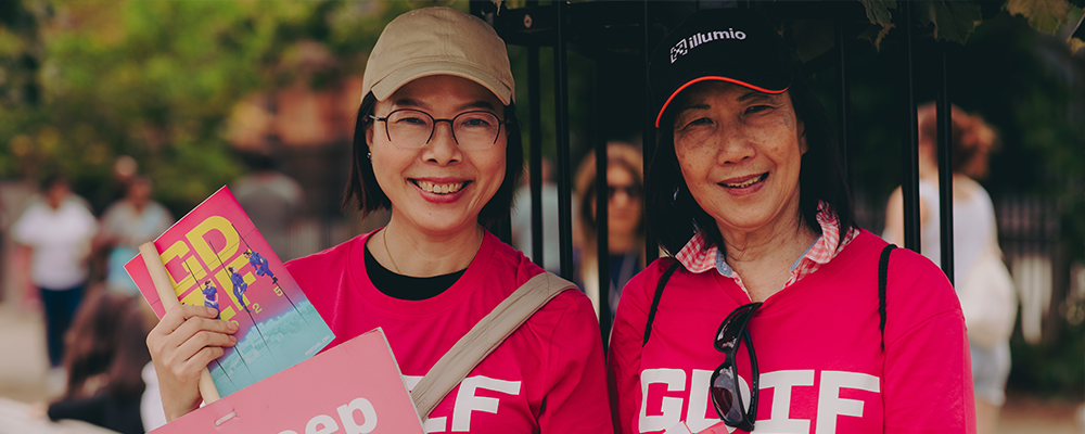Two volunteers smile at the camera outdoors. They wear matching pink T-shirts and caps. One holds leaflets and a booklet. People and trees appear softly blurred in the background.