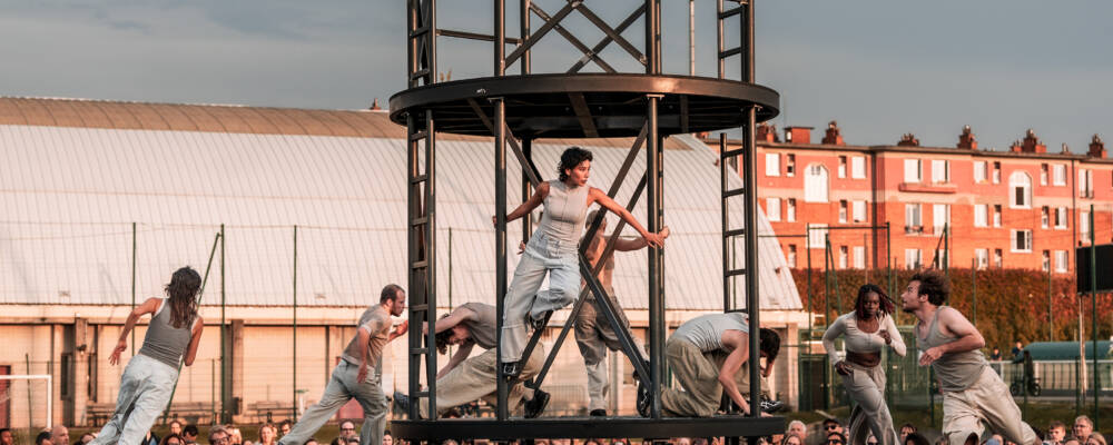 4 dancers dressed in white and grey sprinting around a circular metal structure in the middle of a stage. Within the structure 2 dancers hold on to the railings looking out at the audience that surrounds them. 2 other dancers crouch dramatically either side of them.