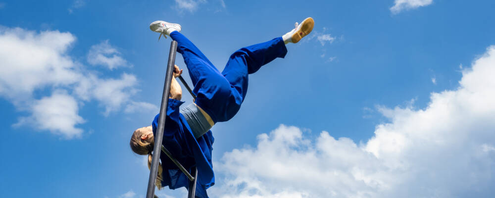 A performer dressed in blue, hangs upside down at the top of a ladder. Behind them a bright blue sky with white fluffy clouds.