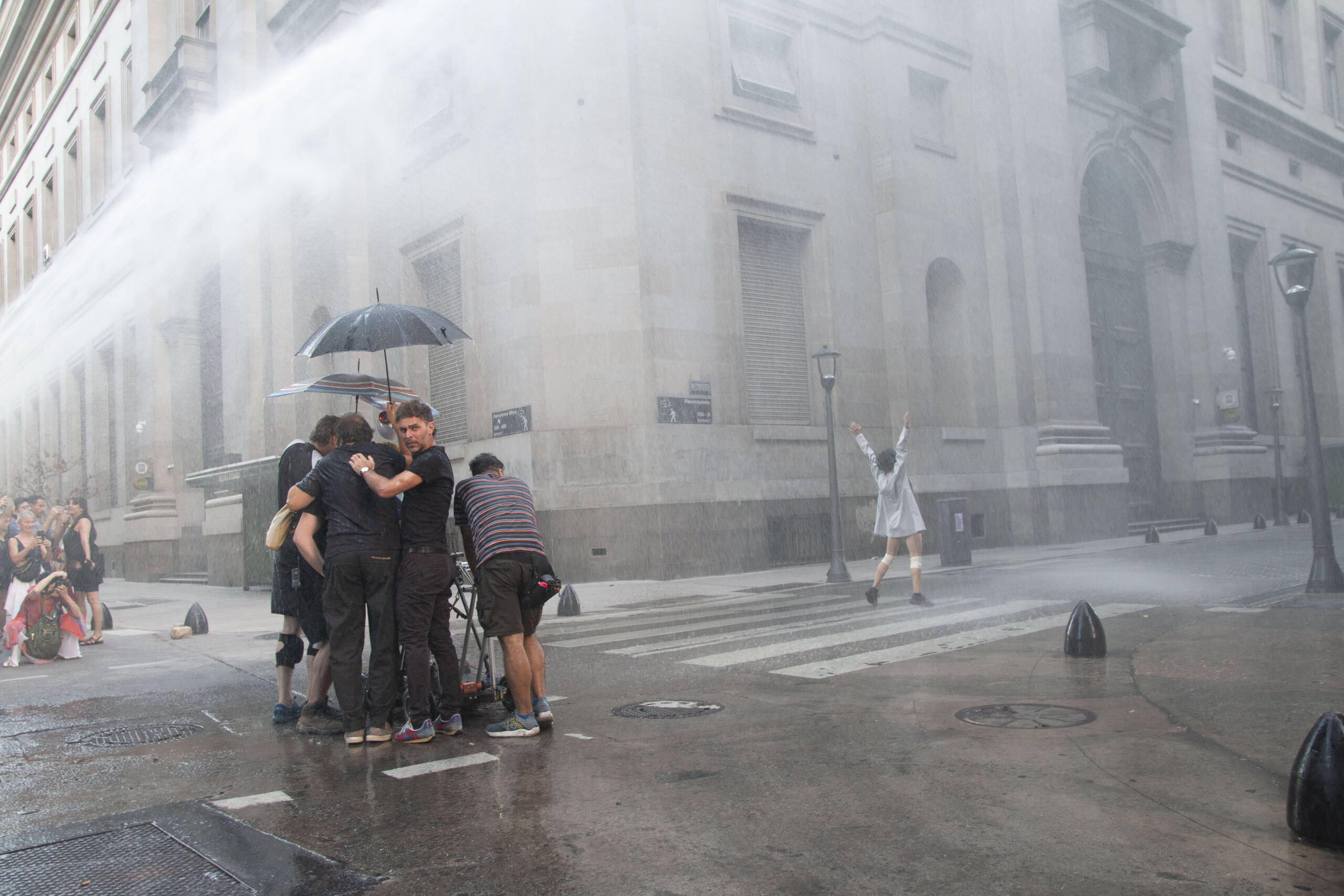 A camera crew huddle under an umbrella in a street as it rains heavily. There is someone in the background dancing joyfully.