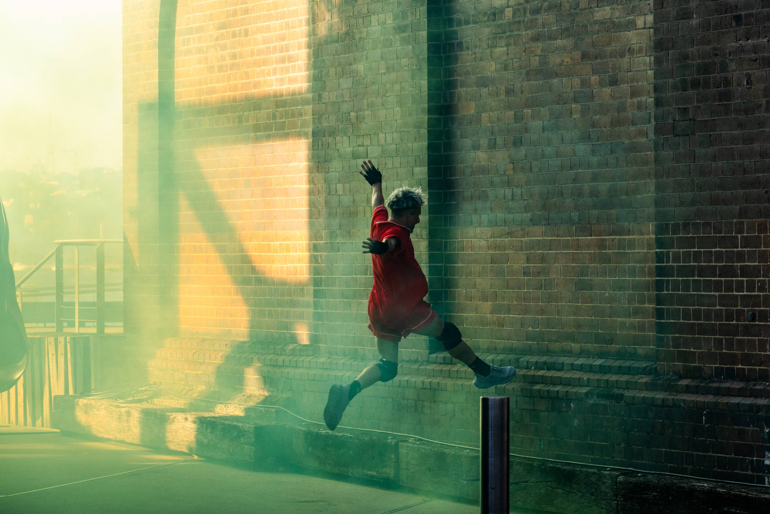 A dancer dressed in red leaps towards a bollard in a street. They are surrounded by green haze.