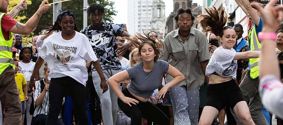 A diverse ensemble of 5 performer dressed in casual clothes dance energetically. They are in front of a busy crowd of people in a town centre and are framed by 2 people in high vis vests.