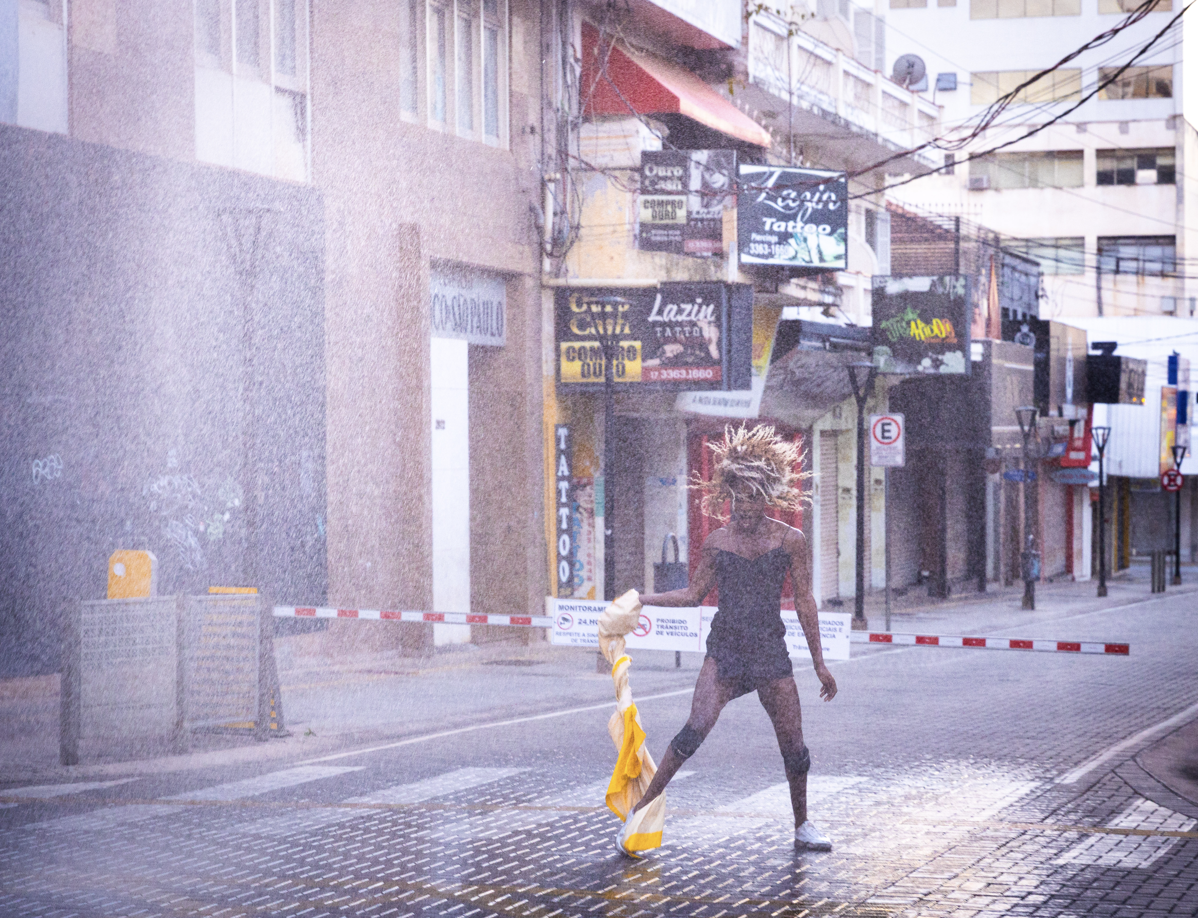 A performer in a mini dress, holding a rain coat in their hand. They are shaking their hair wildly whilst it rains heavily on them.