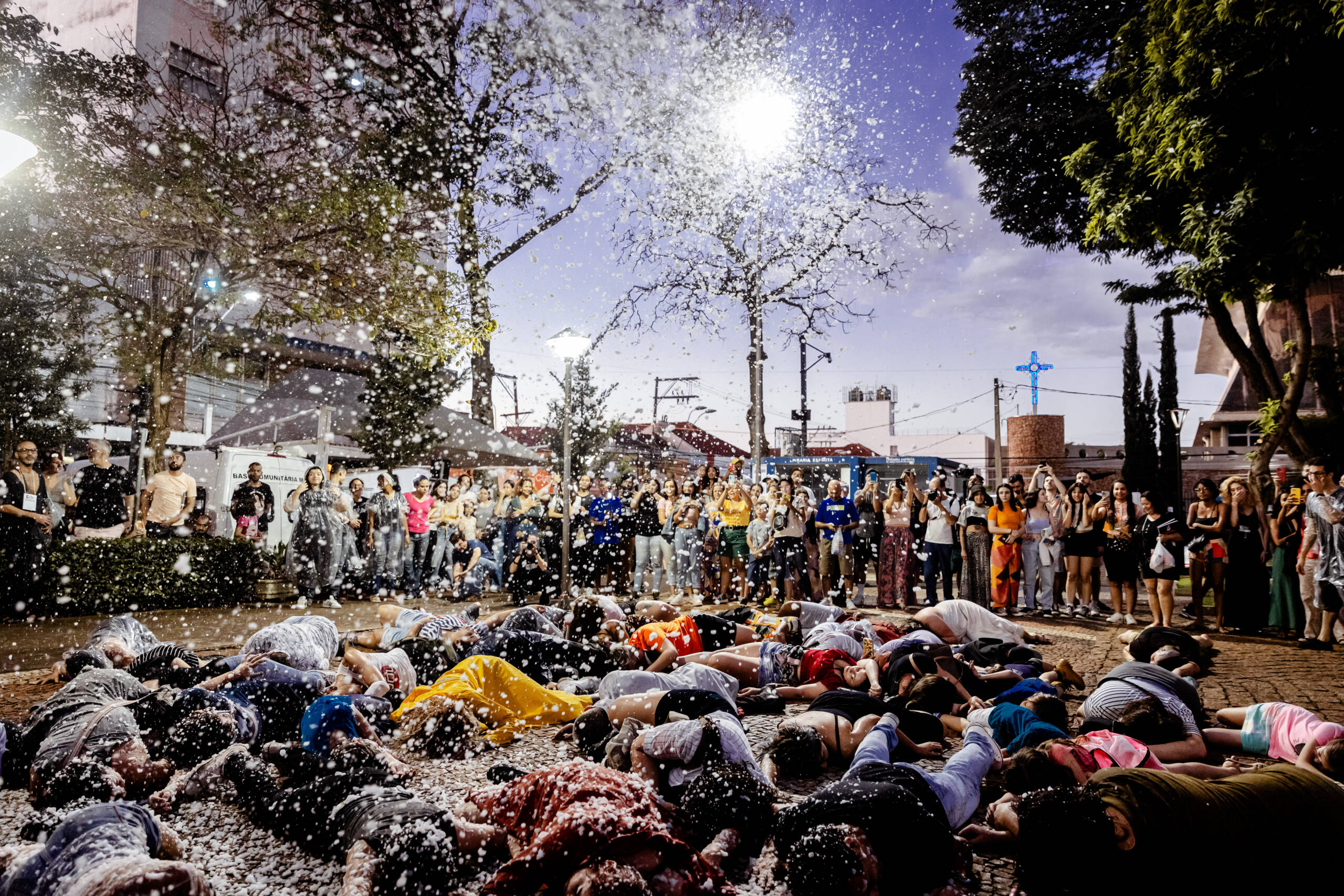 A group of people lying on the floor, covered in snow from a snow machine. They are being watched by an audience.
