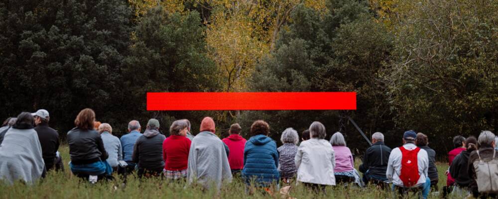 A crowd of people sat on grass staring at an LED screen that glows red. Behind the screen is a forest