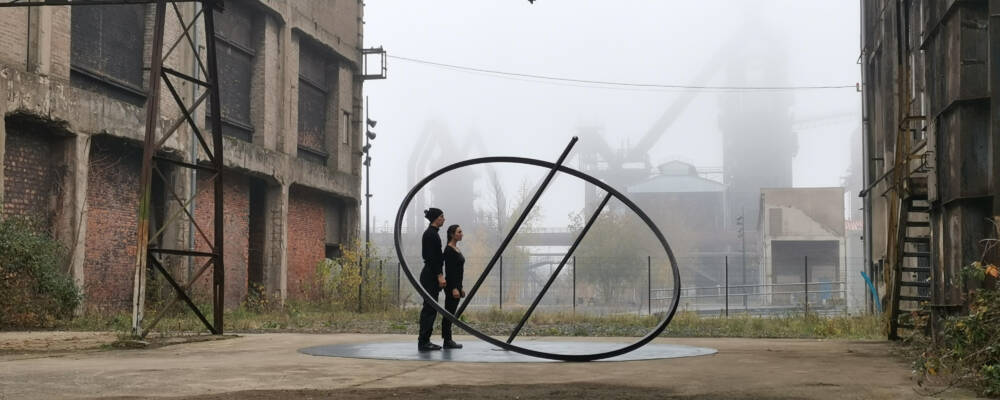 Two figures stand in an industrial space looking up at circular metal structure.